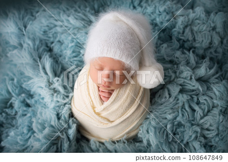 Sleeping newborn baby boy in the first days of life in a white soft cocoon with a knitted woolen white hat on a blue background. Studio macro photography, portrait of a newborn. Woman's happiness.  108647849