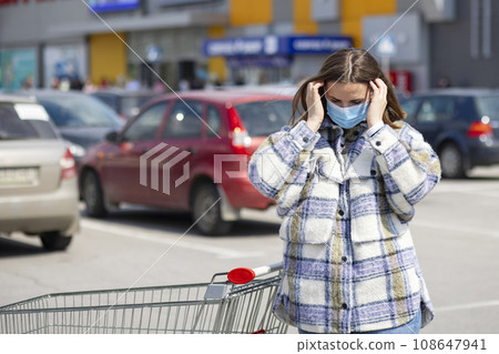 A young woman in a medical mask stands in a parking lot near a grocery store. A young woman in a medical mask stands in a parking lot near a grocery store. 108647941