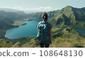 A beautiful brunette traveler in a cyan T-shirt and backpack, looking at a lake in the valley. Mountain forest landscape in summer against the sky with clouds. 108648130