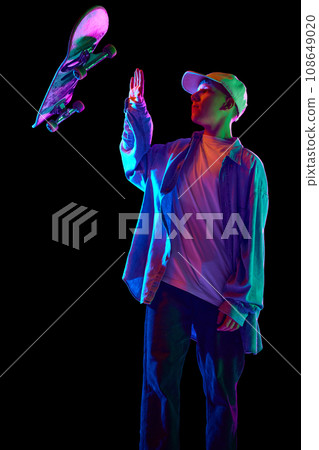 Boy in casual clothes standing with skateboard, posing, doing tricks against black studio background in neon light Boy in casual clothes standing with skateboard, posing, doing tricks against black studio background in neon light 108649020