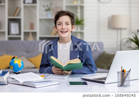 Portrait of a schoolboy at home in the living room, the boy is studying sitting at the desk, reading a book and doing homework, the teenager is smiling and looking at the camera. Portrait of a schoolboy at home in the living room, the boy is studying sitting at the desk, reading a book and doing homework, the teenager is smiling and looking at the camera. 108649248