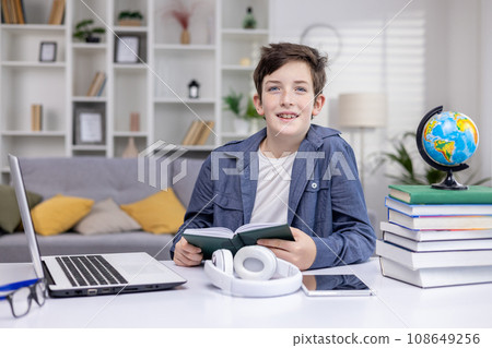 Portrait of a schoolboy at home in the living room, the boy is studying sitting at the desk, reading a book and doing homework, the teenager is smiling and looking at the camera. Portrait of a schoolboy at home in the living room, the boy is studying sitting at the desk, reading a book and doing homework, the teenager is smiling and looking at the camera. 108649256