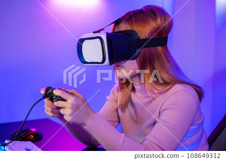 Gamer in VR headset glasses exploring metaverse play video game online with joystick on computer neon lights, Excited woman virtual playing gamepad controller she very determined at home Gamer in VR headset glasses exploring metaverse play video game online with joystick on computer neon lights, Excited woman virtual playing gamepad controller she very determined at home 108649312