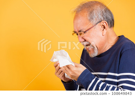 Portrait senior old man blowing nose with tissue studio shot isolated on yellow background, Asian elder man cold having flu and sneezing from sickness virus problem use tissue, pensioner unwell 108649843