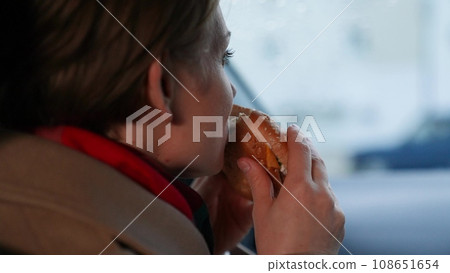 Famished lady takes bite of burger to quell hunger sitting in vehicle on cloudy day. Burger delivered for lady eating burger in vehicle. Lady indulging in unhealthy burger while driving in vehicle 108651654