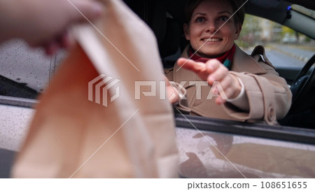 Fast food delivery guy hands offer bag to delighted woman in car with man. Woman receives fast food from delivery guy sitting in car. Order made by delivery guy and woman waiting for fast food in car Fast food delivery guy hands offer bag to delighted woman in car with man. Woman receives fast food from delivery guy sitting in car. Order made by delivery guy and woman waiting for fast food in car 108651655