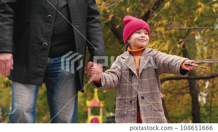 Smiling little girl and father walk joining hands across autumn park. Cheerful little daughter holds father hand walking in fall park. Caring father and little daughter rest in garden on autumn day Smiling little girl and father walk joining hands across autumn park. Cheerful little daughter holds father hand walking in fall park. Caring father and little daughter rest in garden on autumn day 108651666