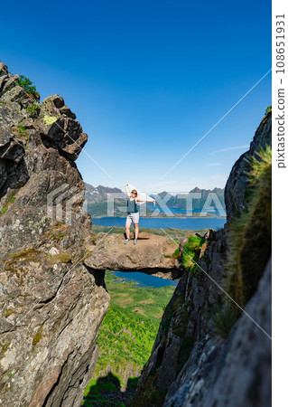 Brave traveler woman standing on hanging stone between rocks. Djevelporten in Norway Lofoten islands Brave traveler woman standing on hanging stone between rocks. Djevelporten in Norway Lofoten islands 108651931