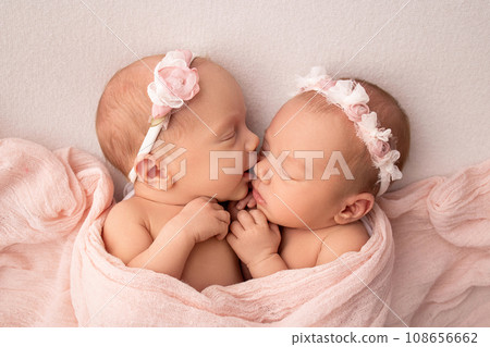 Tiny newborn twin girls. A newborn twin sleeps next to his sister. Newborn twin girls on the background of a pink blanket with pink bandages. The girls gently hug and kiss their sister in a cute pose 108656662