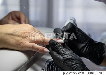 Manicurist at work. Professional manicurist removes old worn nail design from nails of client using modern electric drill. Preparing fingernails for making fresh manicure and applying new nail design 108656706