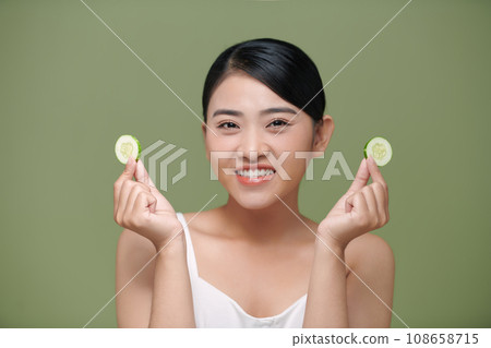 Young beautiful asian lady holding slices of cucumber near her face and smiling Young beautiful asian lady holding slices of cucumber near her face and smiling 108658715