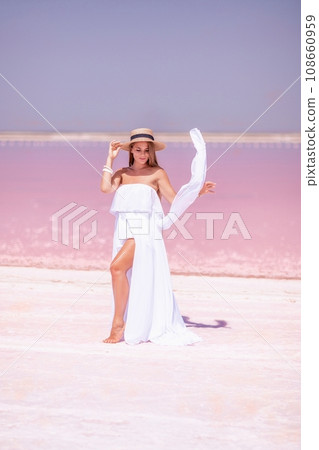 Woman in pink salt lake. She in a white dress and hat enjoys the scenic view of a pink salt lake as she walks along the white, salty shore, creating a lasting memory. Woman in pink salt lake. She in a white dress and hat enjoys the scenic view of a pink salt lake as she walks along the white, salty shore, creating a lasting memory. 108660959