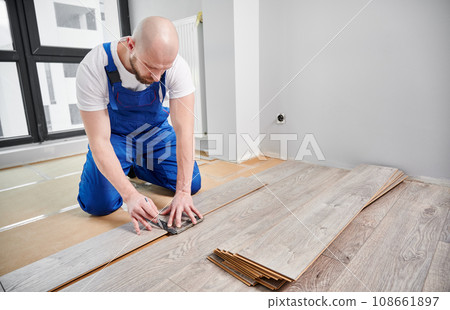 Man in work overalls using metal construction ruler and pen while drawing line on laminate flooring board. Male floor installer preparing laminate plank for installation in apartment under renovation. Man in work overalls using metal construction ruler and pen while drawing line on laminate flooring board. Male floor installer preparing laminate plank for installation in apartment under renovation. 108661897