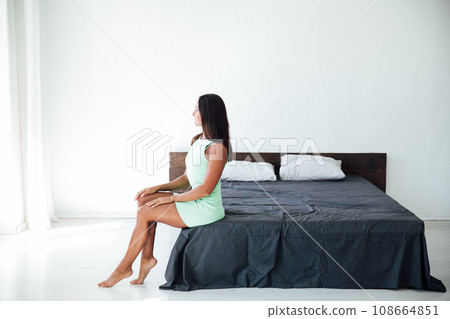 Brunette woman sitting on bed in bright room in bedroom Brunette woman sitting on bed in bright room in bedroom 108664851