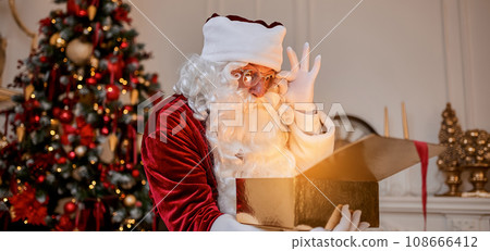 Surprised Santa Claus with glowing gift near christmas tree. New year and Merry Christmas concept 108666412