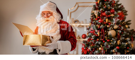 Surprised Santa Claus with glowing gift near christmas tree. New year and Merry Christmas concept Surprised Santa Claus with glowing gift near christmas tree. New year and Merry Christmas concept 108666414