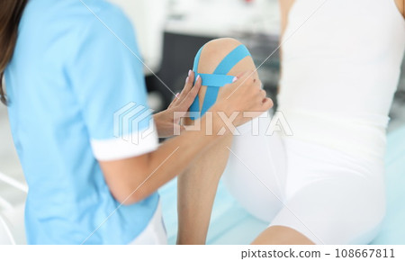 Rehabilitation therapist gluing blue tapes on woman knee closeup Rehabilitation therapist gluing blue tapes on woman knee closeup 108667811