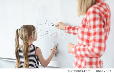 Little girl and teacher solving math equations on blackboard 108667883