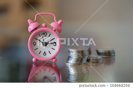Pink alarm clock standing near pile of coins closeup Pink alarm clock standing near pile of coins closeup 108668208