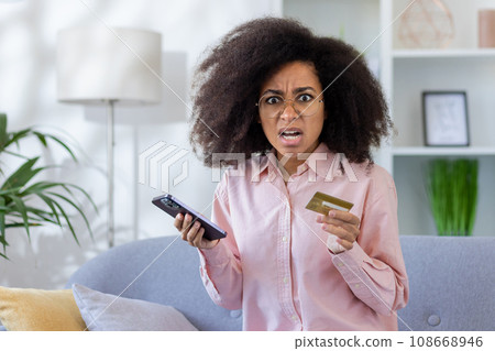 Shocked cheated woman at home sitting on sofa in living room, online buyer looking at camera, scared hispanic holding phone, bank credit card in hands, received money transfer error, payment refused. 108668946