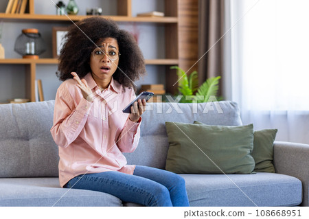 Upset disappointed woman at home received online message message with bad news, depressed hispanic woman sitting on sofa in living room looking at camera, holding phone. 108668951