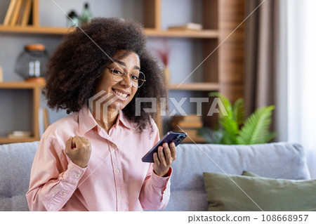 Portrait of happy confident woman indoors, sitting on sofa in living room smiling and looking at camera, good news. Winner holding phone in hands, satisfied result, achievement celebrating. Portrait of happy confident woman indoors, sitting on sofa in living room smiling and looking at camera, good news. Winner holding phone in hands, satisfied result, achievement celebrating. 108668957