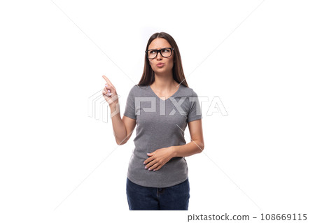 well-groomed young woman in glasses dressed in a gray t-shirt points to the space for advertising 108669115