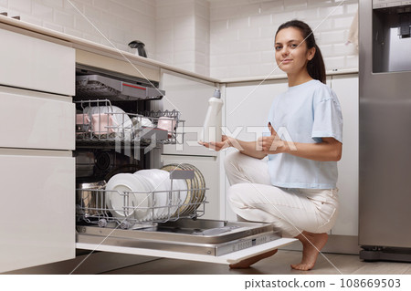 woman pours rinse aid into the dishwasher compartment in modern white kitchen 108669503