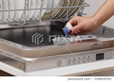 female hand puts dishwasher tablet into open automatic built-in dishwasher machine with dirty dish inside in kitchen 108669504