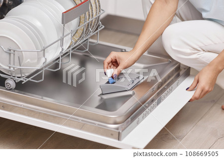female hand puts dishwasher tablet into open automatic built-in dishwasher machine with dirty dish inside in kitchen 108669505
