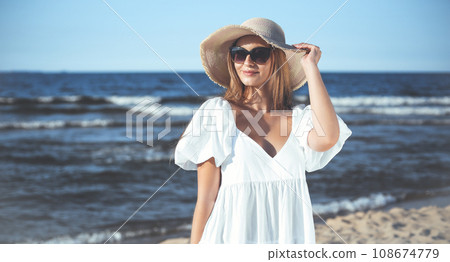 Happy blonde woman is posing on the ocean beach with sunglasses and a hat. Evening sun 108674779