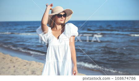 Happy smiling blonde woman is posing on the ocean beach with sunglasses and a hat 108674786