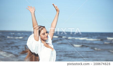 Happy smiling beautiful woman on the ocean beach standing in a white summer dress, raising hands Happy smiling beautiful woman on the ocean beach standing in a white summer dress, raising hands 108674828