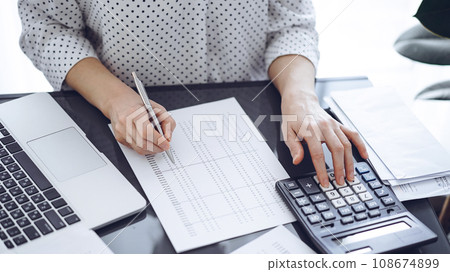 Woman accountant using a calculator and laptop computer while counting taxes with a client or a colleague. Business audit team, finance advisor Woman accountant using a calculator and laptop computer while counting taxes with a client or a colleague. Business audit team, finance advisor 108674899