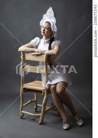 Portrait of a beautiful girl in a snow maiden costume on a gray background. A Russian snow girl in a crown sits on a wooden chair. 108675345