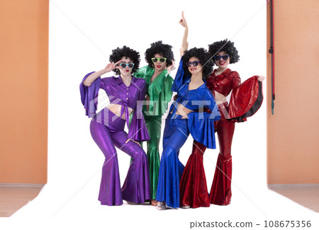 A group of girls in colorful flared suits and afro wigs pose in a photo studio. Disco style from the eighties or seventies. 108675356