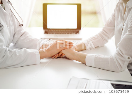 Doctor and patient sitting at the white table near window in clinic. Female physician's hands reassuring woman. Medicine concept 108675439