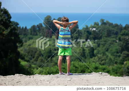 The boy stands with his hands folded in front of his head and admires the beautiful seascape. 108675716