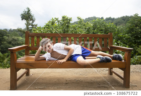 A happy child in the summer heat lies on a wooden bench against the backdrop of tropical plants. The boy is on vacation in the summer. A happy child in the summer heat lies on a wooden bench against the backdrop of tropical plants. The boy is on vacation in the summer. 108675722