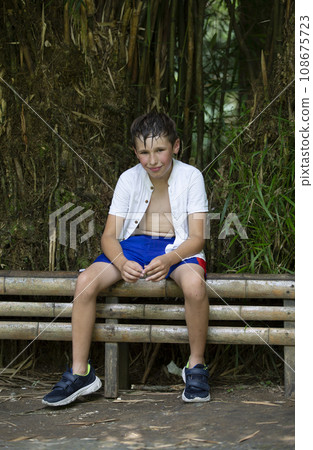 A happy child sits on a bench in the summer heat against the backdrop of tropical plants. A happy child sits on a bench in the summer heat against the backdrop of tropical plants. 108675723