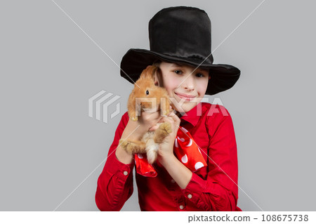 A magician boy in a black hat is holding a red rabbit. 108675738