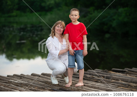 Happy mother and son stand by the river on a wooden bridge, pose and look at the camera. 108676118