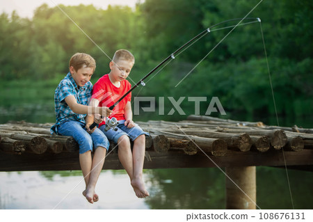 Two boy friends are sitting on a wooden bridge and fishing. Children on a fishing trip. Two boy friends are sitting on a wooden bridge and fishing. Children on a fishing trip. 108676131