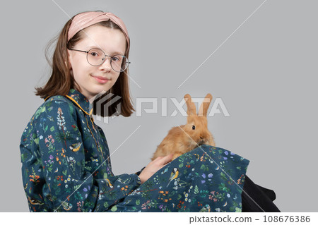 Beautiful little girl in glasses plays with a red rabbit on a gray background. 108676386