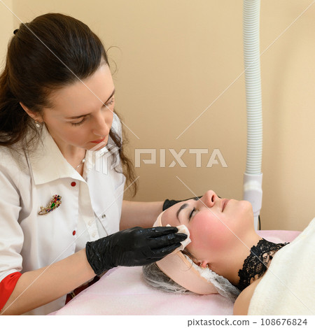 The cosmetologist uses a napkin to remove the remains of the cream from the face. 108676824