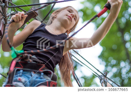 Teenage girl hiking in the rope park in safety equipment. Safety harness. Teenage girl hiking in the rope park in safety equipment. Safety harness. 108677665