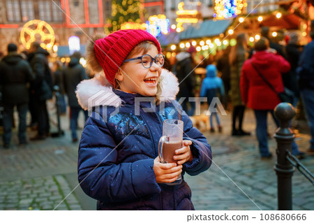 Little cute preschool girl drinking hot children punch or chocolate on German Christmas market. Happy child on traditional family market in Germany, Laughing kid in colorful winter clothes 108680666