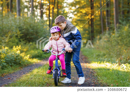 Cute little preschool girl in safety helmet riding bicycle. School kid boy, brother teaching happy healthy sister child cycling and having fun with learning bike. Active siblings family outdoors. 108680672