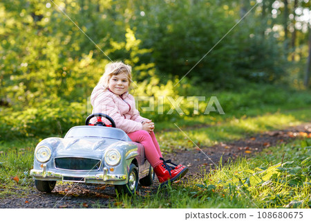 Little preschool girl driving big vintage toy car. Happy child having fun with playing outdoors. Active preschooler child enjoying warm autumn day in forest. Smiling stunning kid playing 108680675