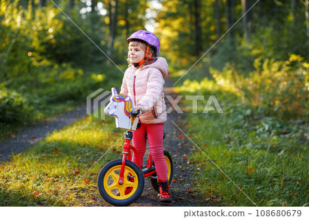 Child riding balance bike. Kids on bicycle in sunny forest. Little girl enjoying to ride glider bike on warm day. Preschooler learning to balance on run bicycle in safe helmet. Sport activity Child riding balance bike. Kids on bicycle in sunny forest. Little girl enjoying to ride glider bike on warm day. Preschooler learning to balance on run bicycle in safe helmet. Sport activity 108680679